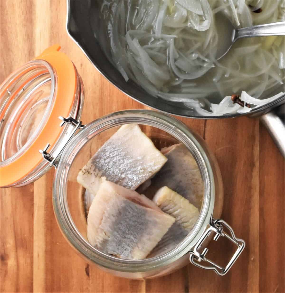 Pieces of herring in open jar and onion vinegar mixture in saucepan.