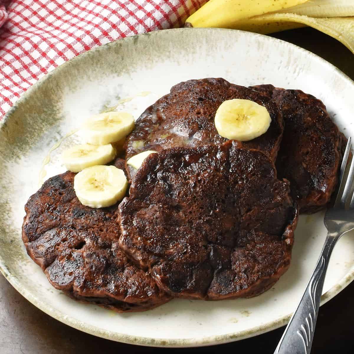 Close-up side view of banana chocolate pancakes with sliced banana and fork on plate.