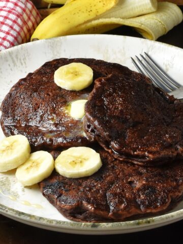 Side view of 3 chocolate banana pancakes with sliced banana on white plate with fork.