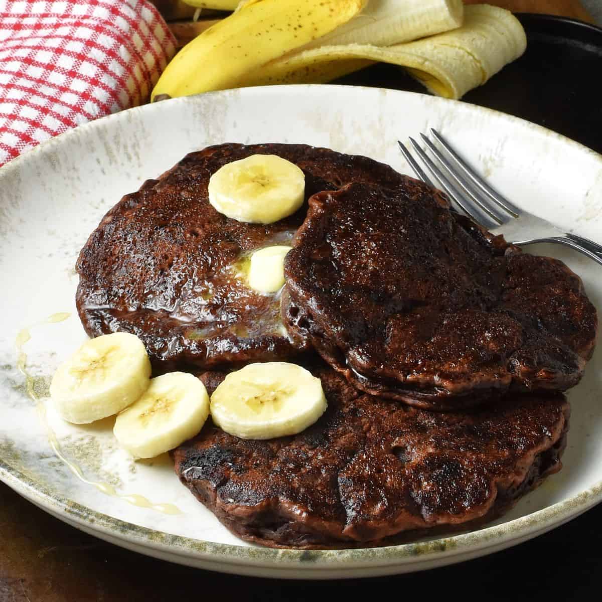 Side view of 3 chocolate banana pancakes with sliced banana on white plate with fork.