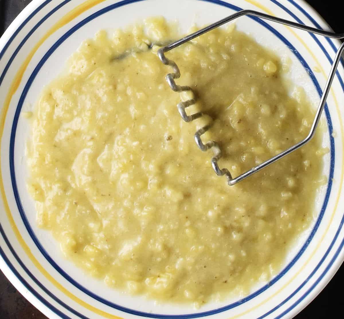 Top down view of mashed banana in large shallow bowl and potato masher.