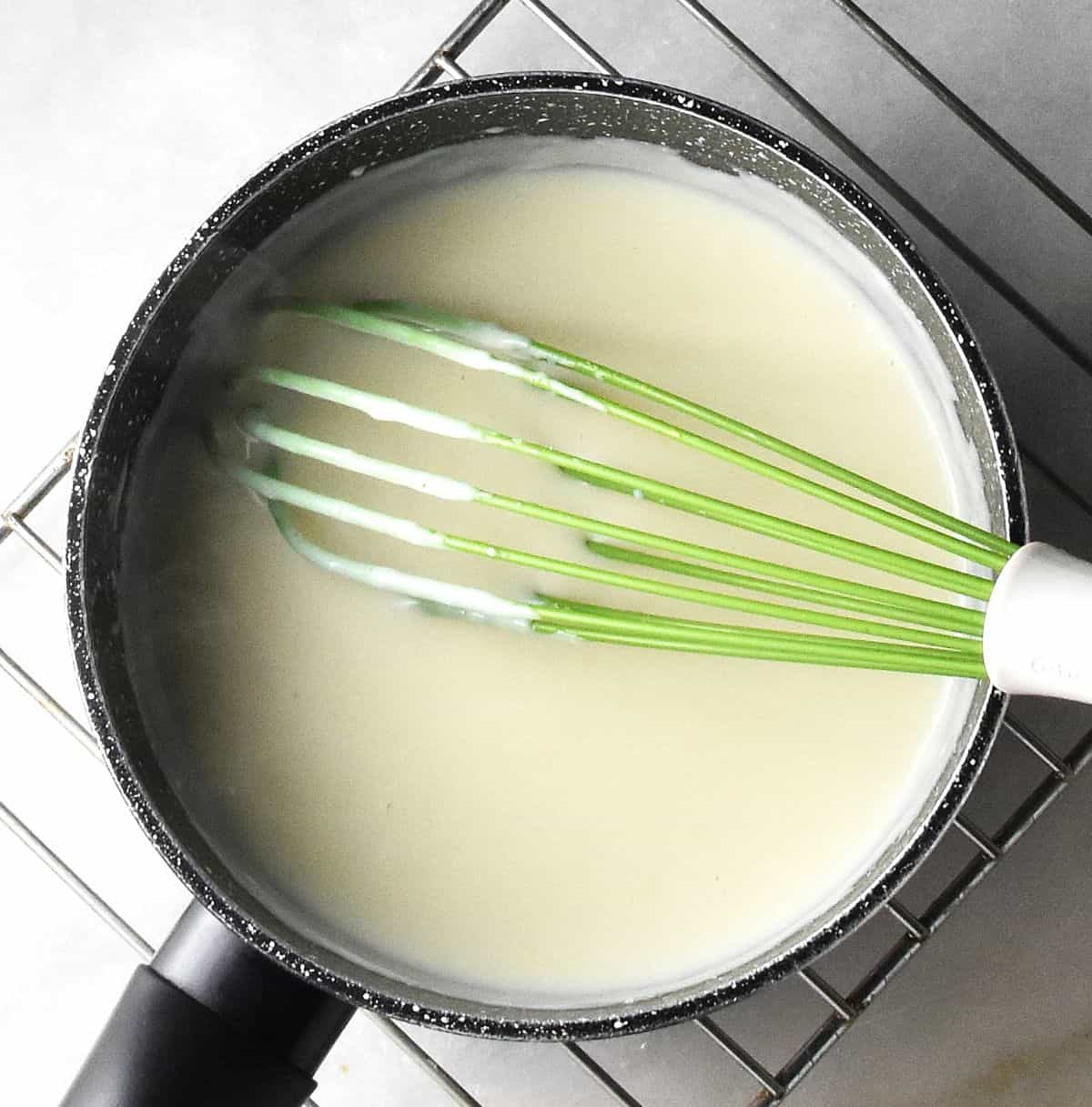 Top down view of creamy bechamel sauce in pot with green whisk.