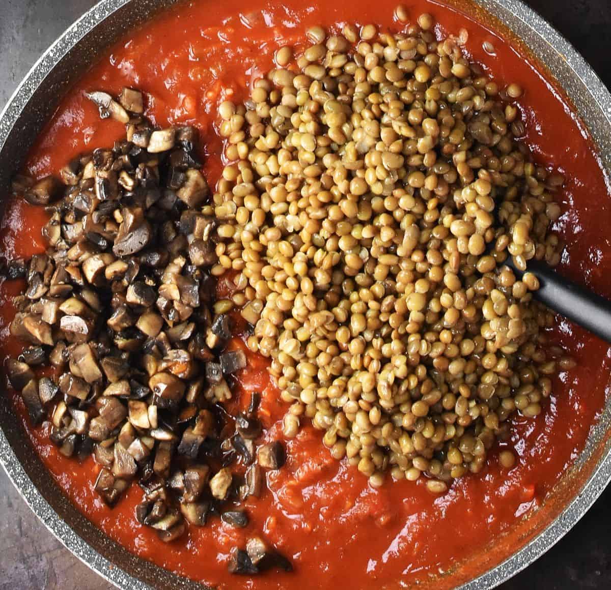 Brown lentils and chopped mushrooms on top of tomato sauce in large pan.