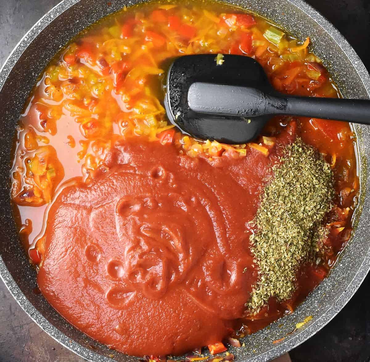 Chopped vegetables with passata and herbs on top in large pan with black spoon.