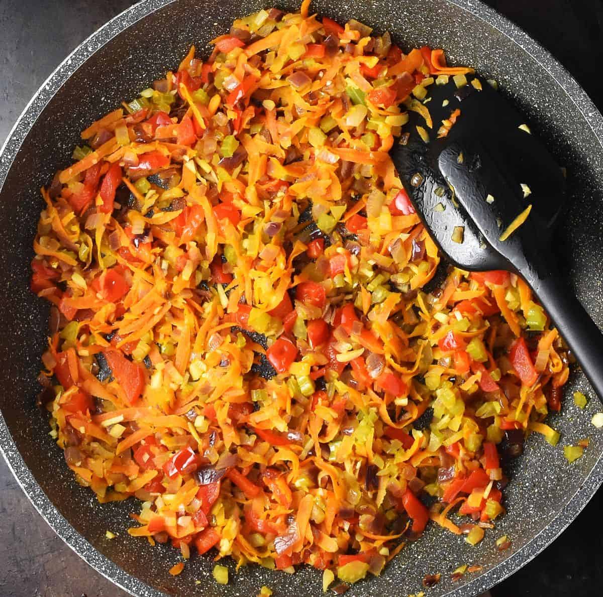 Finely chopped vegetables cooking in large, shallow pan with black silicone spatula.