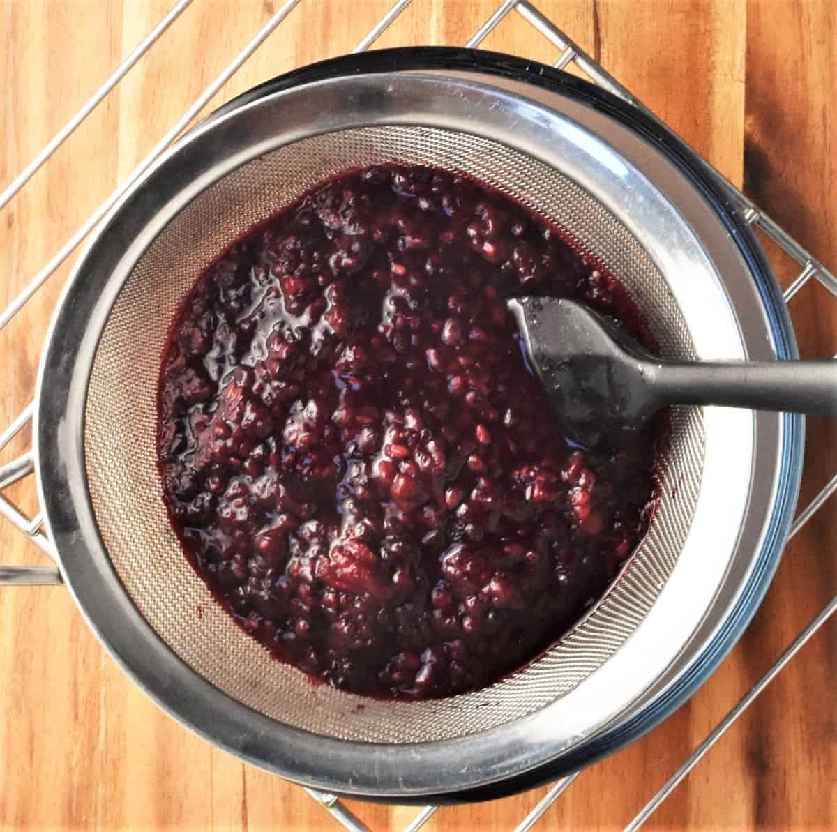 Cooked blackberries in sieve with black spatula separating seeds from juice.