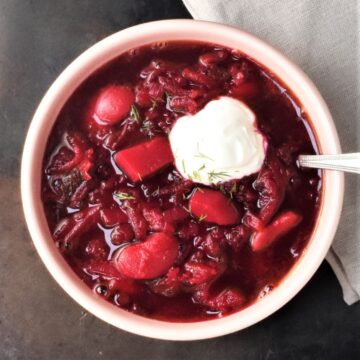 Top down view of vegetarian borscht with sour cream and spoon in pink bowl.