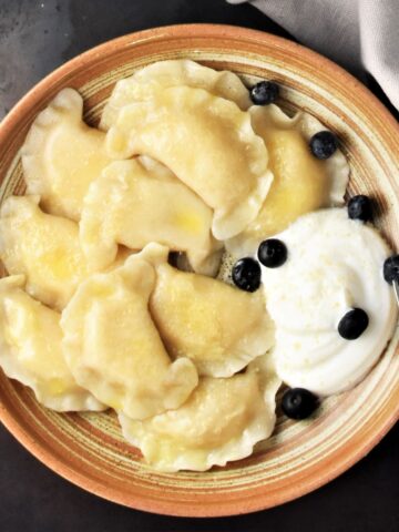 Top down view of Polish sweet cheese pierogi in brown bowl with sour cream and fruit.