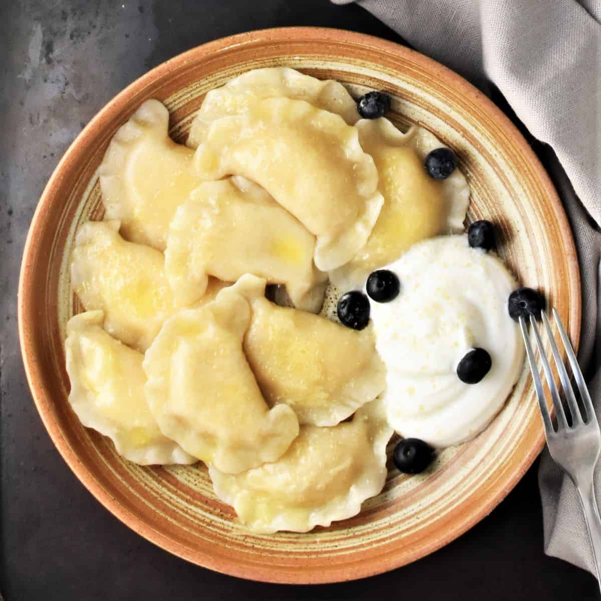 Top down view of Polish sweet cheese pierogi in brown bowl with sour cream and fruit.