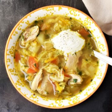 Top down view of chicken cabbage soup in yellow bowl with dollop of sauerkraut and spoon.