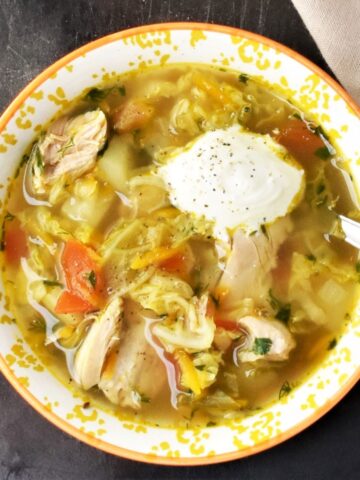 Top down view of chicken cabbage soup in yellow bowl with dollop of sauerkraut and spoon.