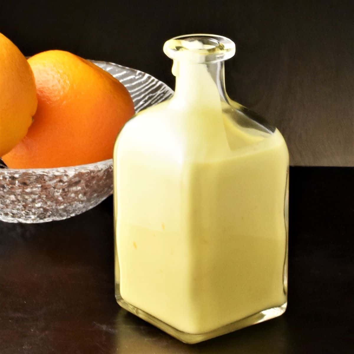 Side view of creamy orange salad dressing in bottle and oranges in background.