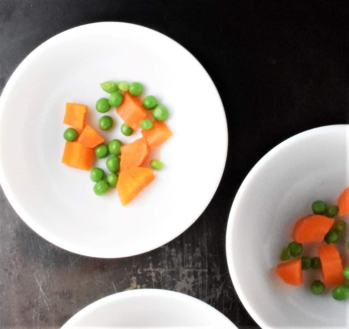 Chopped carrots and peas in white bowls.