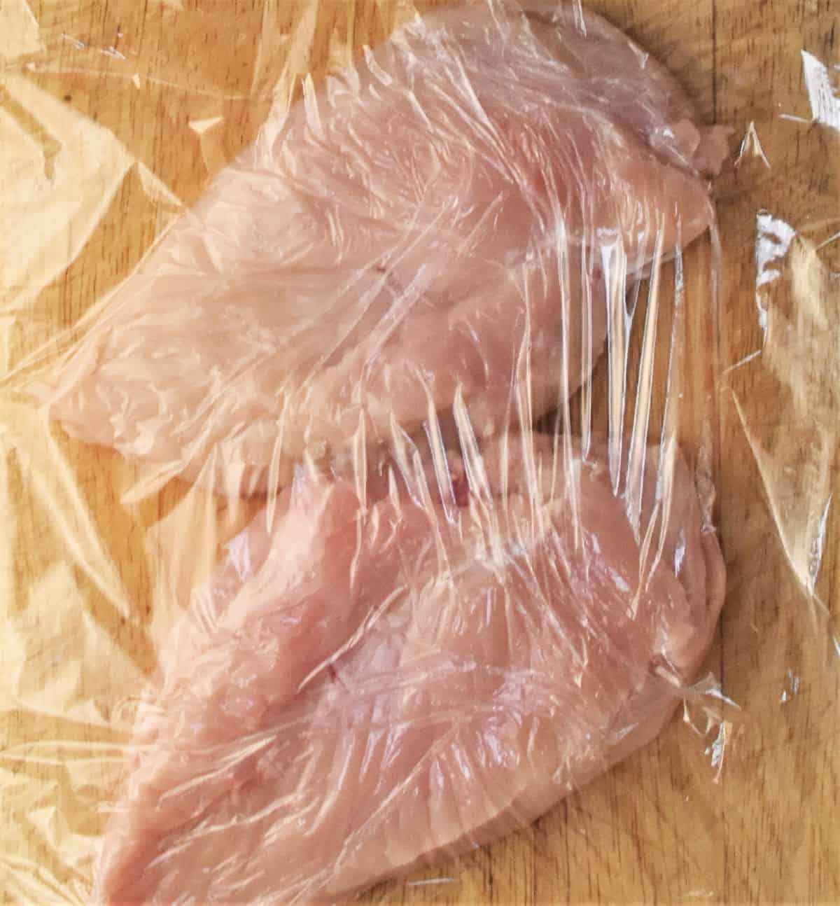 2 chicken breasts covered with plastic wrap on cutting board.