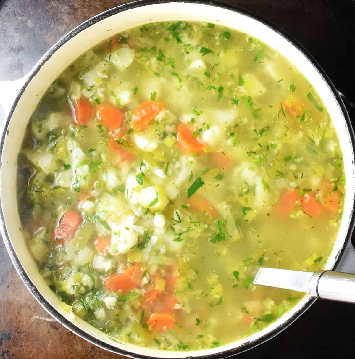 Top down view of soup with chunks of cauliflower, potatoes and carrots in white pot with ladle.
