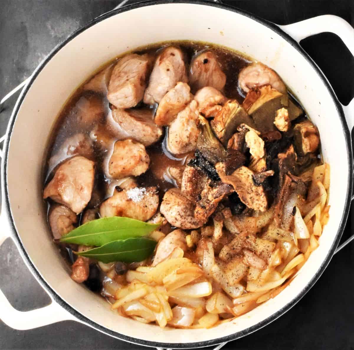 Chunks of pork, sliced onion, bay leaf, mushrooms and water in white pot.
