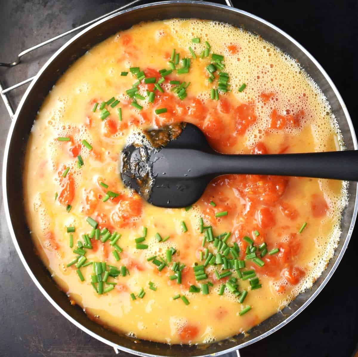 Egg mixture with tomatoes and chives in pan with spatula.