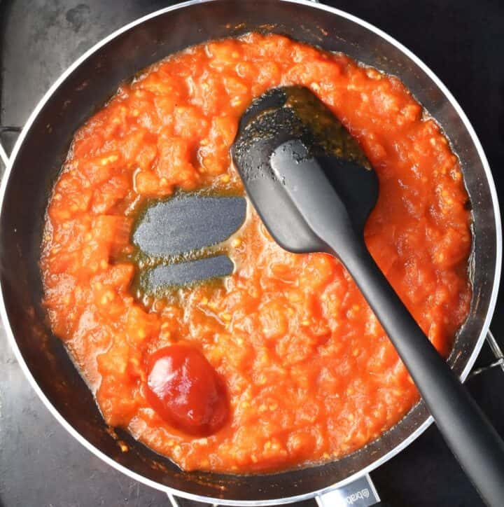 Thick tomato mixture with ketchup and black spatula in pan.