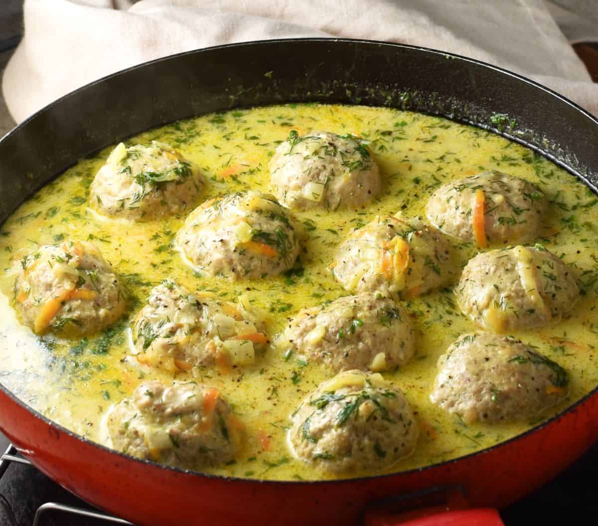 Side view of Polish pulpety meatballs in creamy broth in red shallow pan.