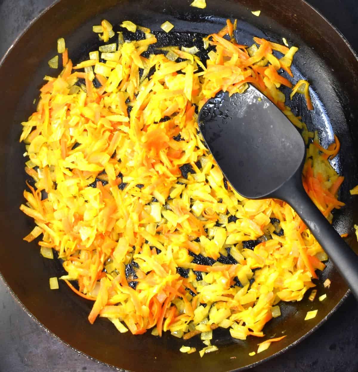 Cooking shredded carrot and onion in pan with black spatula.