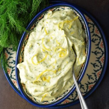 Dill mayonnaise in oval blue bowl with spoon and fresh dill in top left corner.