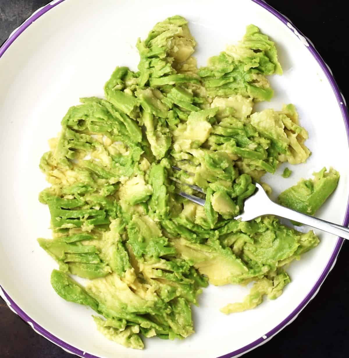 Top down view of mashed avocado in shallow white bowl with fork.