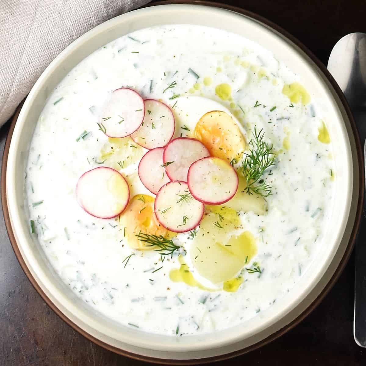 Top down view of creamy cold cucumber soup with radish, eggs and potatoes in grey bowl.
