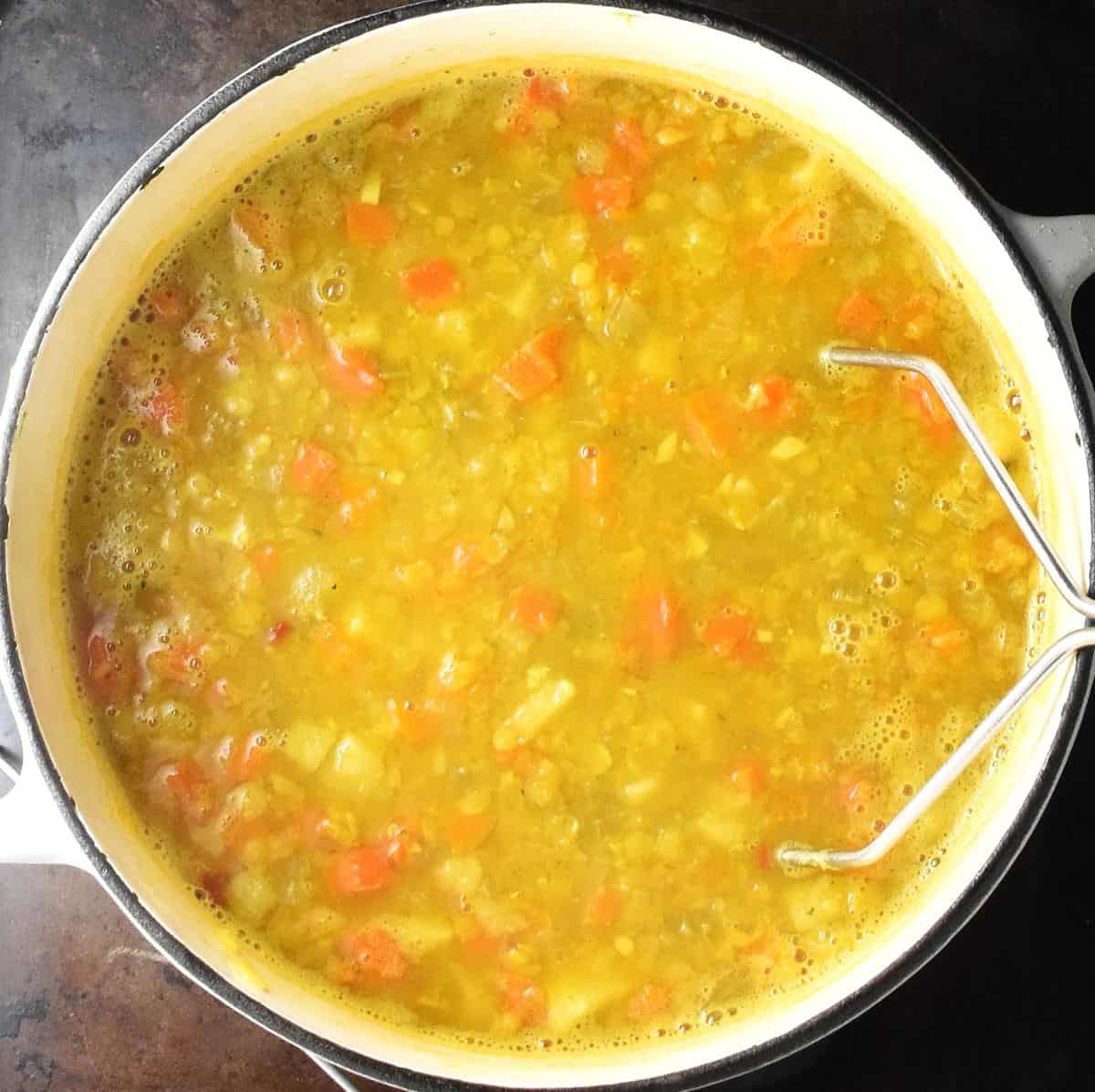 Chunky vegan mulligatawny in large white pot with potato masher.