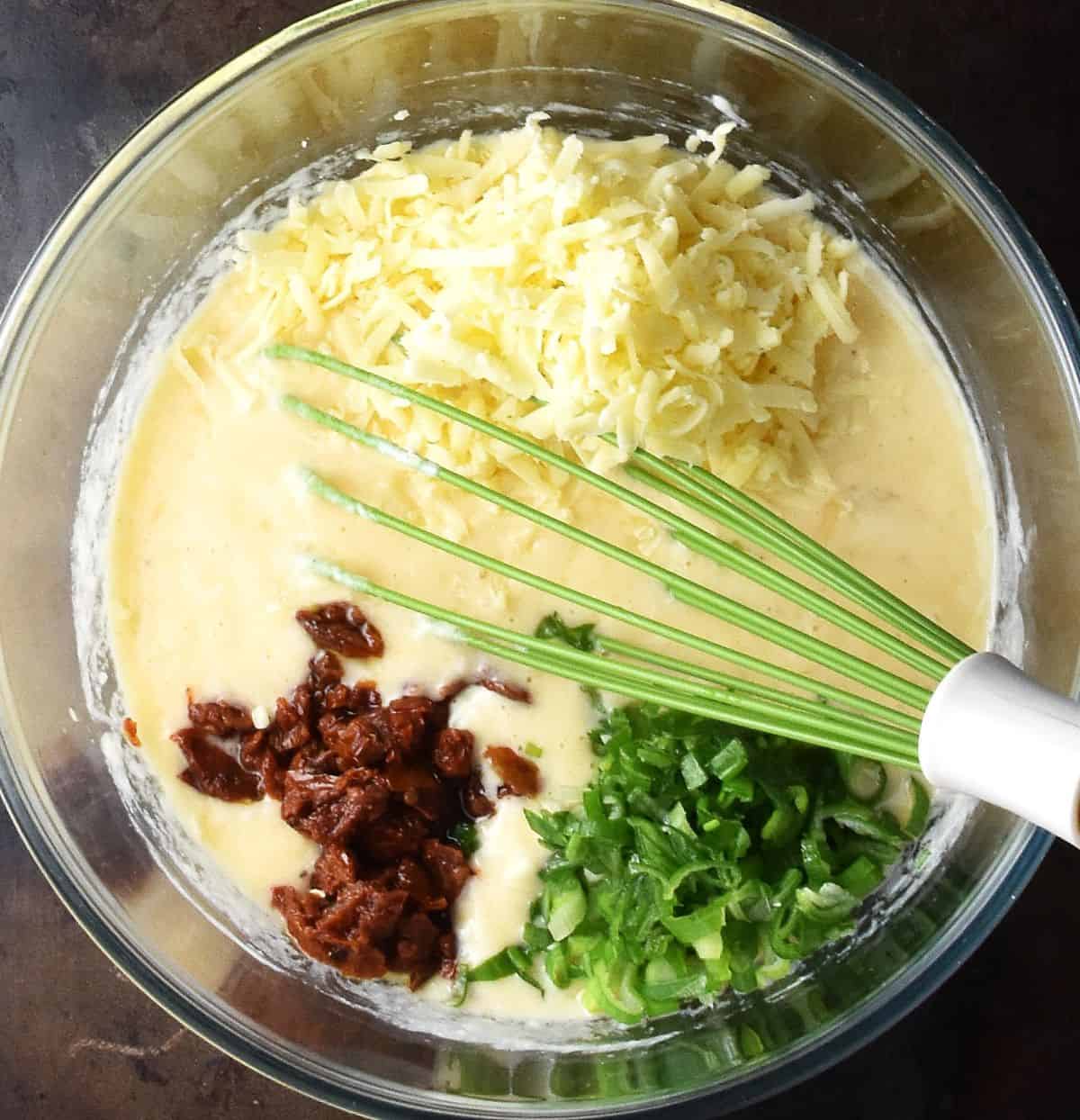 Egg and potato mixture with grated cheese and chopped vegetables in glass bowl with whisk.