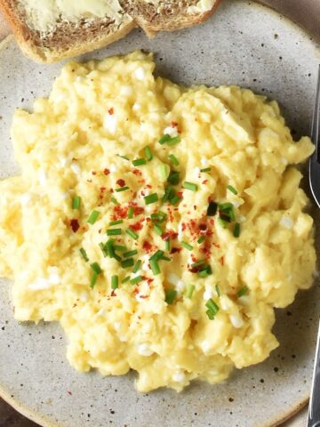 Top down view of creamy scrambled eggs with chives on spotted grey plate with fork.