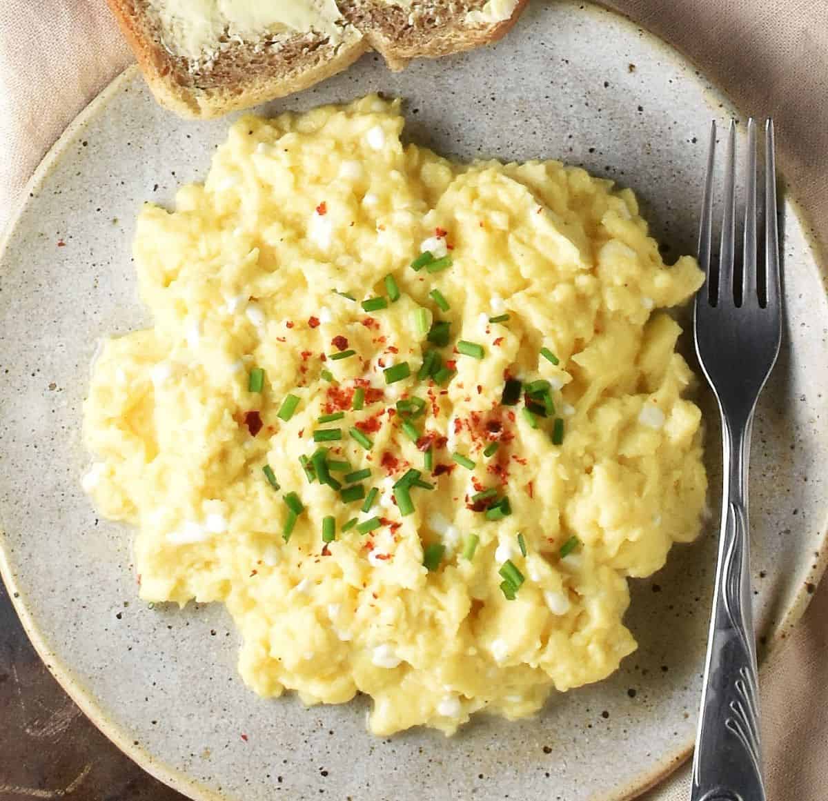 Top down view of creamy scrambled eggs with chives on spotted grey plate with fork.