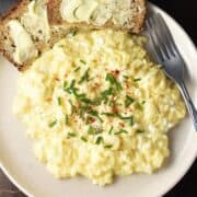 Top down view of creamy cottage cheese scrambled eggs garnished with chives on pink plate with buttered bread.