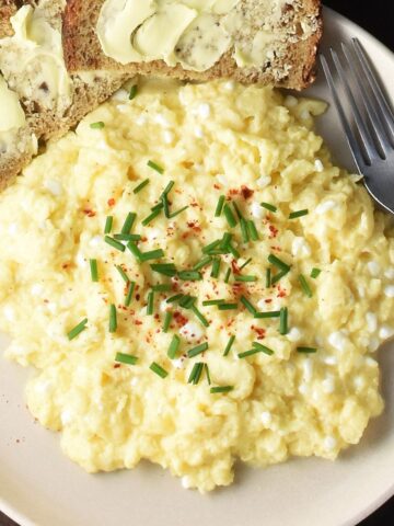 Top down view of creamy cottage cheese scrambled eggs garnished with chives on pink plate with buttered bread.