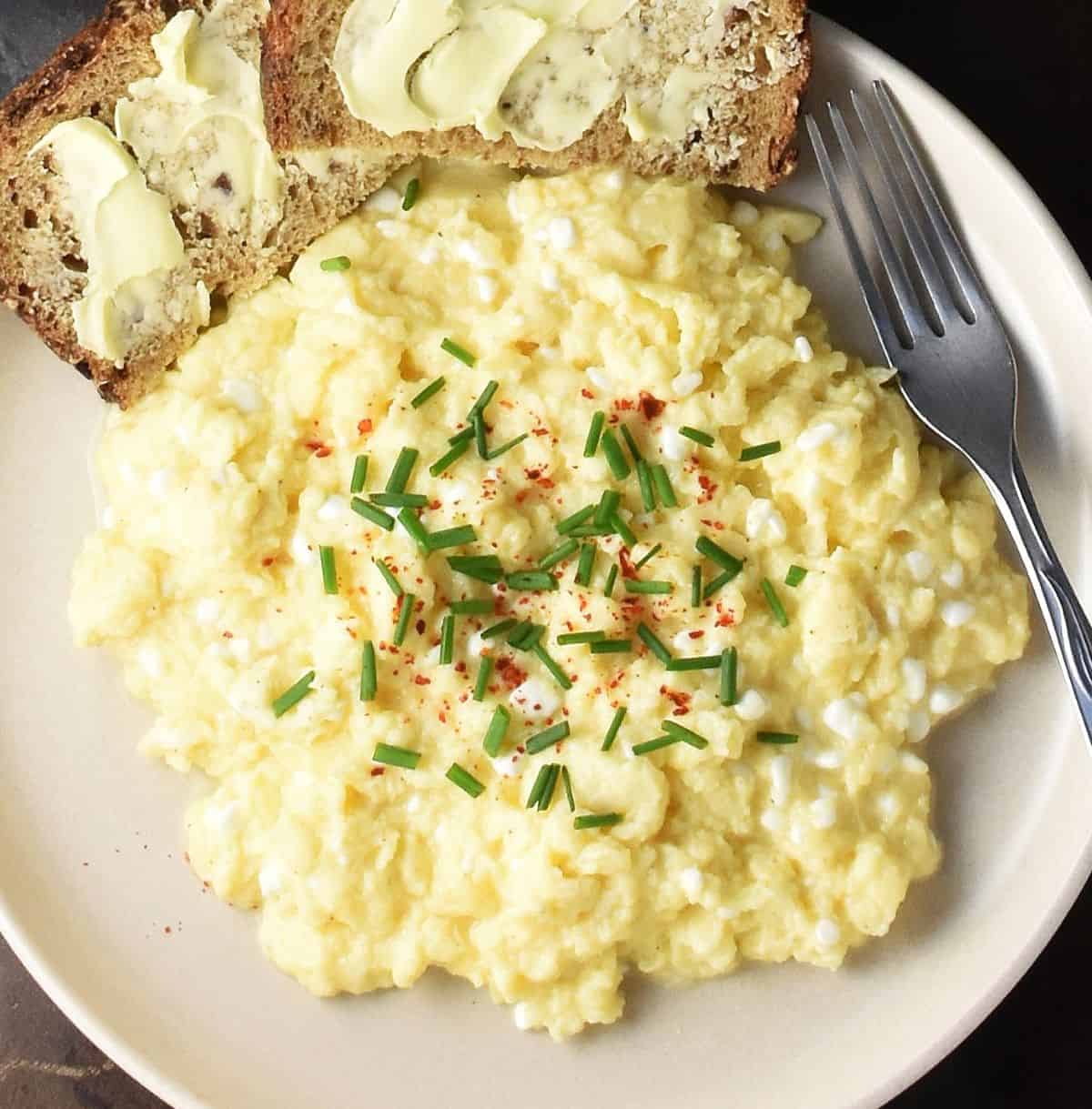 Top down view of creamy cottage cheese scrambled eggs garnished with chives on pink plate with buttered bread.