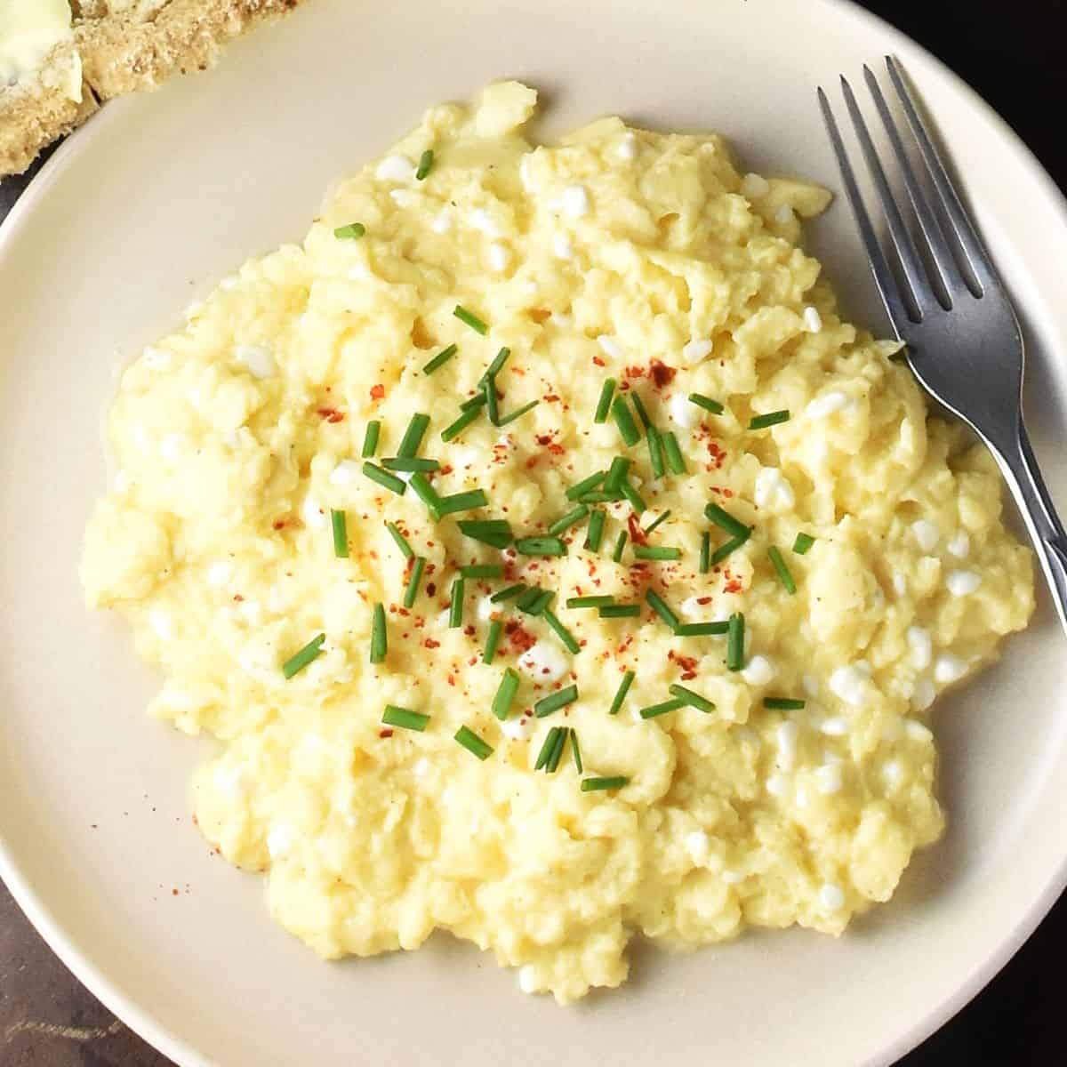 Top down view of scrambled eggs with cottage cheese and chives on pink plate with fork.