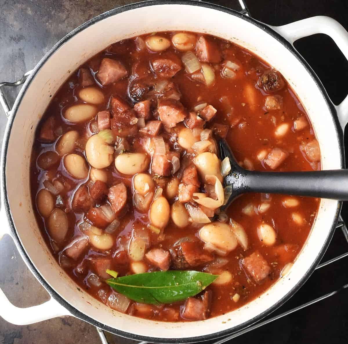 Cooking bean stew with tomato and sausage in white pot with black spatula.