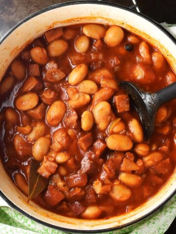 Top down view of butter bean stew in tomato sauce in white pot with black spoon.
