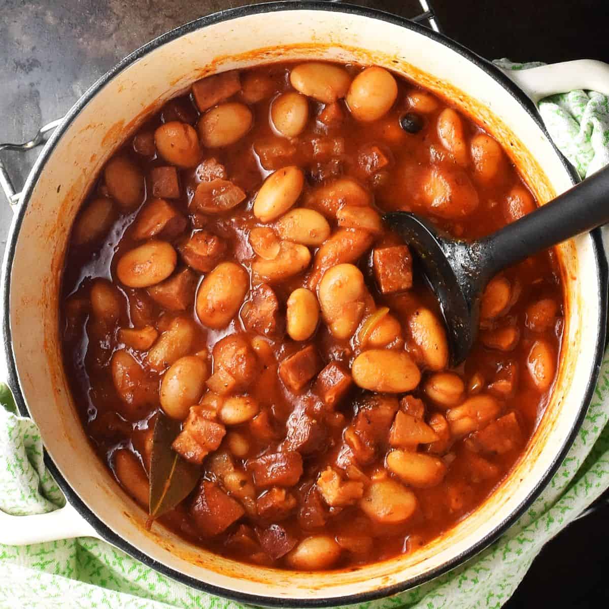 Top down view of butter bean stew in tomato sauce in white pot with black spoon.