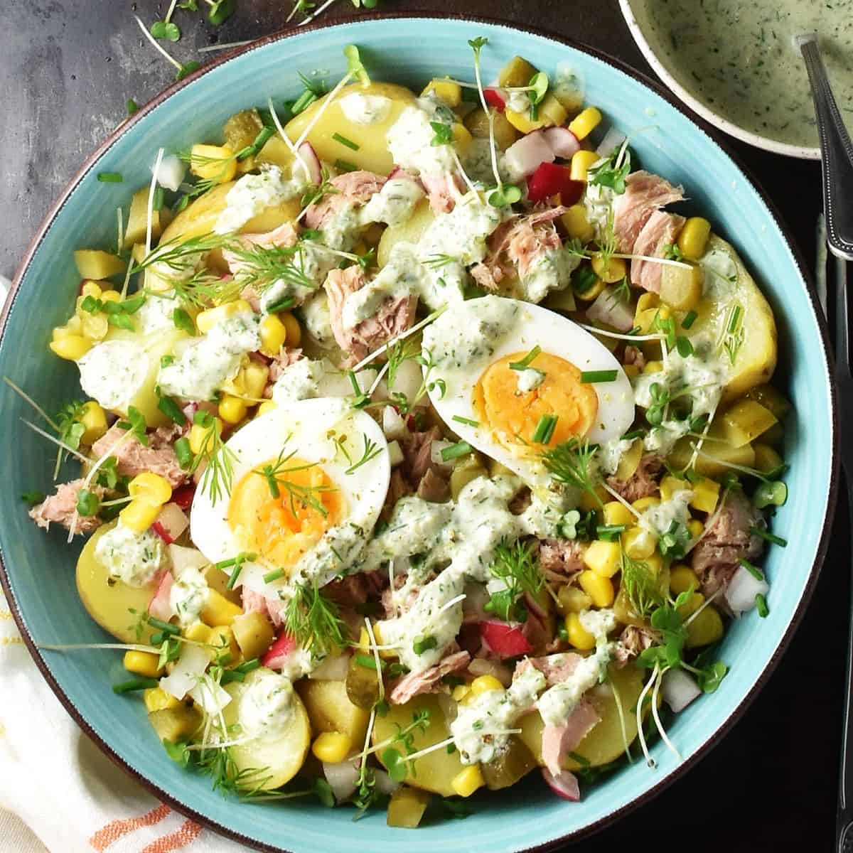 Top down view of potato tuna salad with eggs, herbs and creamy dressing in blue bowl.