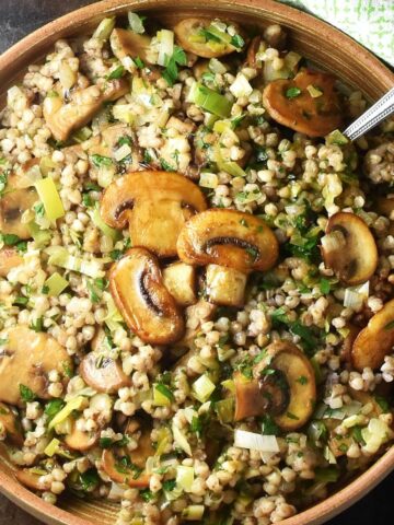 Top down view of cooked buckwheat with browned mushrooms in brown round dish with spoon.