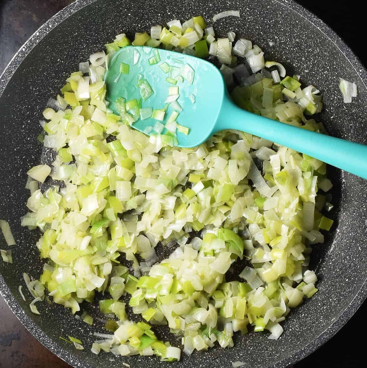Sauteed onion and leek in large pan with blue spatula.
