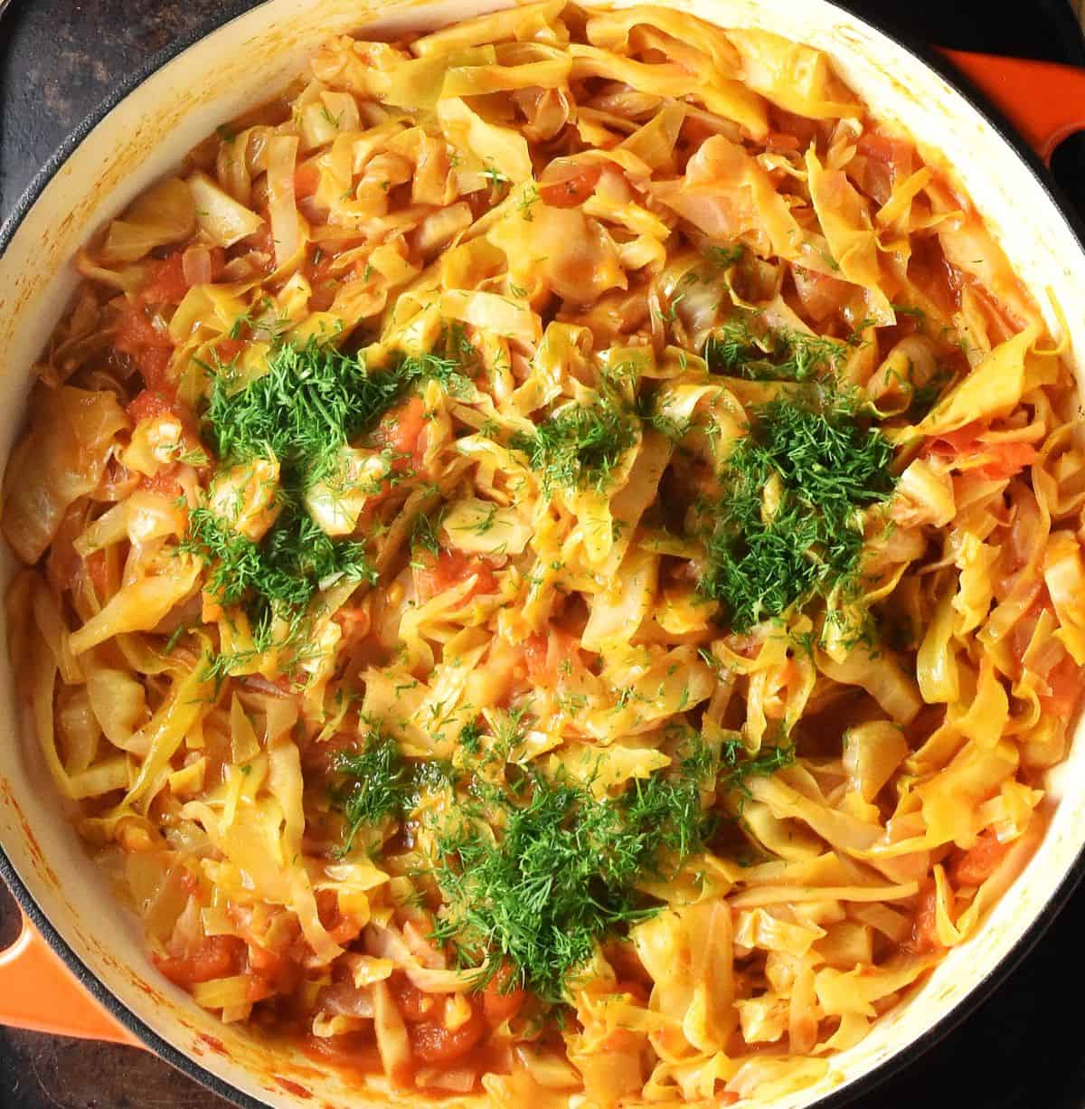 Top down view of cooked cabbage with tomatoes and dill in large white pan.