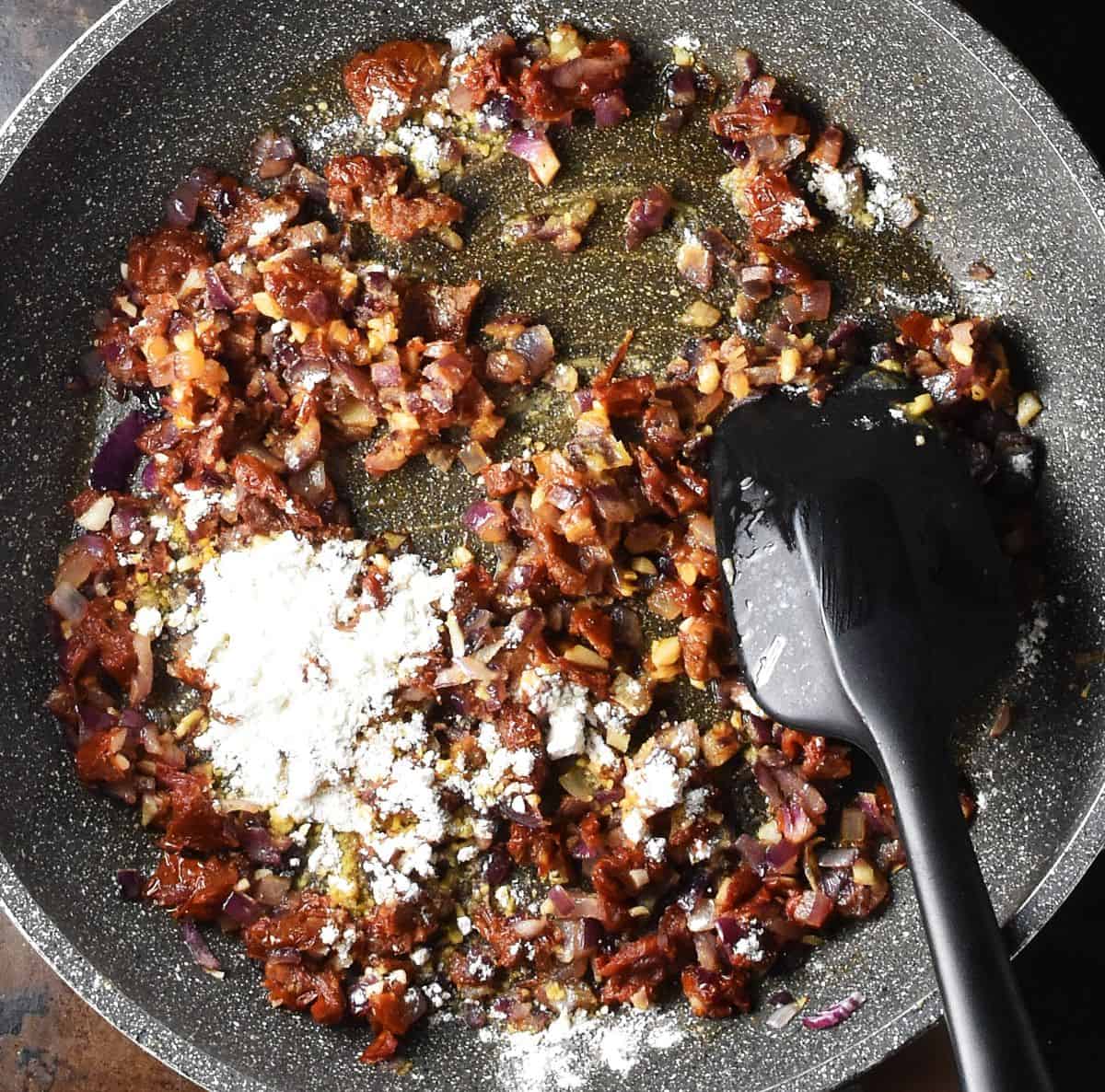Top down view of chopped sundried tomatoes and flour in large pan with black spatula.