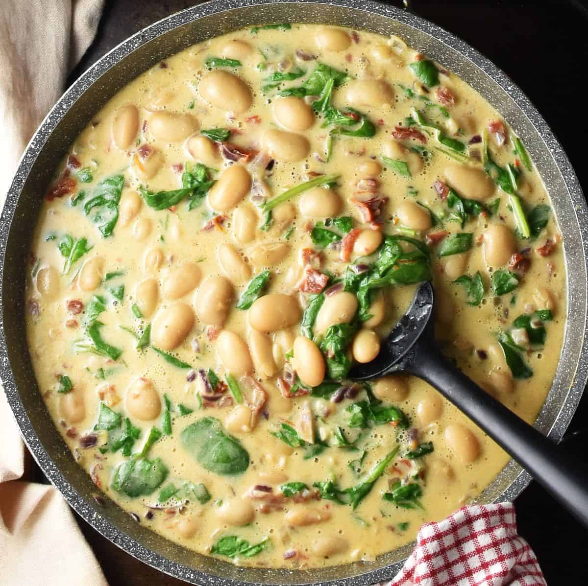 Top down view of butter beans in creamy sauce with spinach in large pan with black spatula.