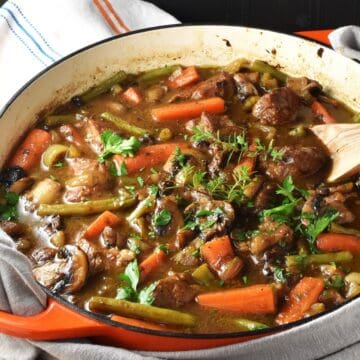 Side view of pork shoulder stew with vegetables and herbs in shallow pan with wooden spoon.