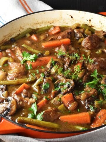 Side view of pork shoulder stew with vegetables and herbs in shallow pan with wooden spoon.