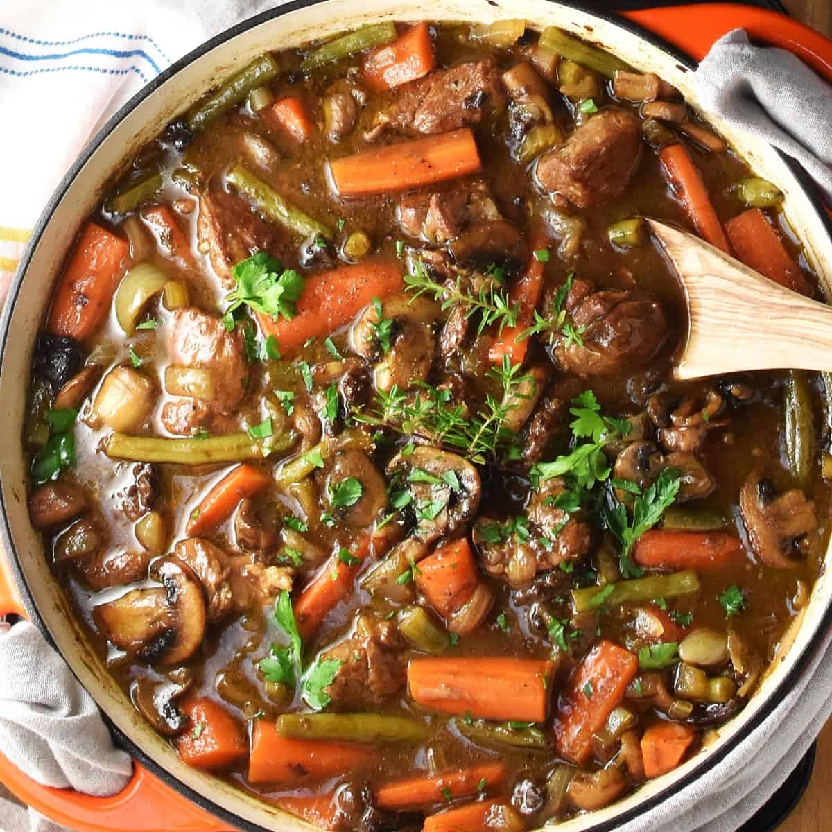 Top down view of stew with pork, vegetables and herbs in shallow pan with wooden spoon.